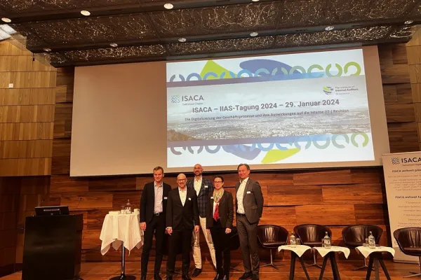 Gruppe von fünf Personen steht auf einer Bühne vor einer Leinwand mit dem Titel der ISACA-IIAS-Tagung 2024 in Zürich. Im Hintergrund sind Stühle und ein Tisch mit Wasserflaschen zu sehen.