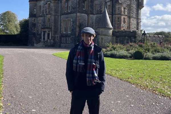 Person mit Mütze und Schal steht auf einer Auffahrt vor einem historischen Schloss aus Stein unter blauem Himmel.