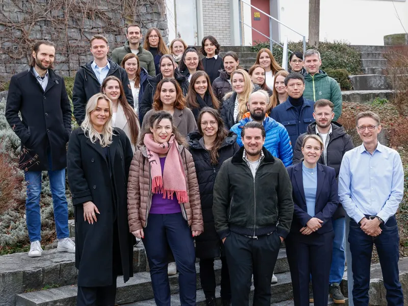 Gruppenfoto von Studierenden und Dozenten des Zertifikatsstudiengangs Compliance-Officer an der Universität Liechtenstein, aufgenommen auf einer Außentreppe. Die Gruppe lächelt in die Kamera, umgeben von winterlicher Vegetation.