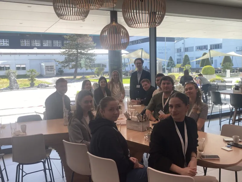 Studierende der Universität Liechtenstein sitzen in einer modernen Cafeteria bei Ivoclar in Schaan. Sie sind um einen Tisch versammelt und blicken in die Kamera. Im Hintergrund sind große Fenster mit Blick auf den Außenbereich zu sehen.